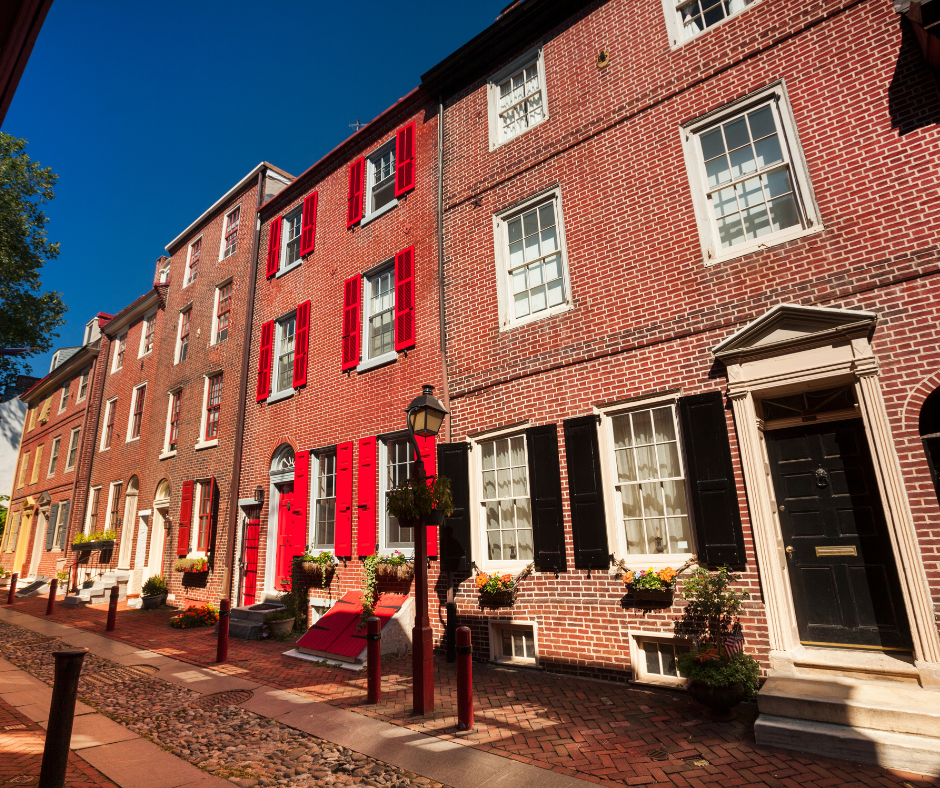 Charming Historic Street with Red-Accent Houses A picturesque view of a historic street featuring brick houses with red shutters and black doors, lined with flower pots and a clear blue sky.